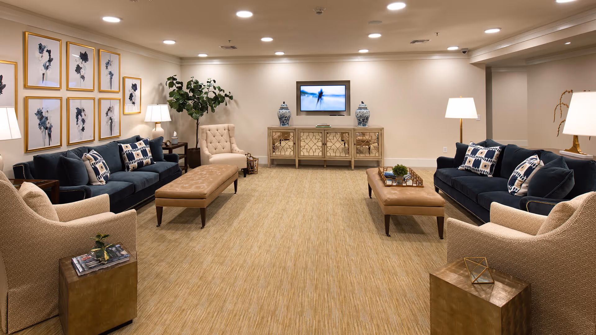 Spacious, well-lit communal lounge with sofas, armchairs, ottomans, lamps, a wall-mounted TV and framed artwork.