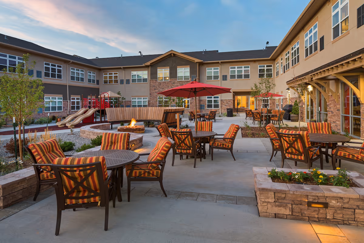 Outdoor courtyard area of Melody Living facility with multiple round tables and chairs featuring striped cushions, red umbrellas, a fire pit with a curved wooden bench, a small playground with slides, and surrounding two-story building under a partly cloudy sky at dusk.