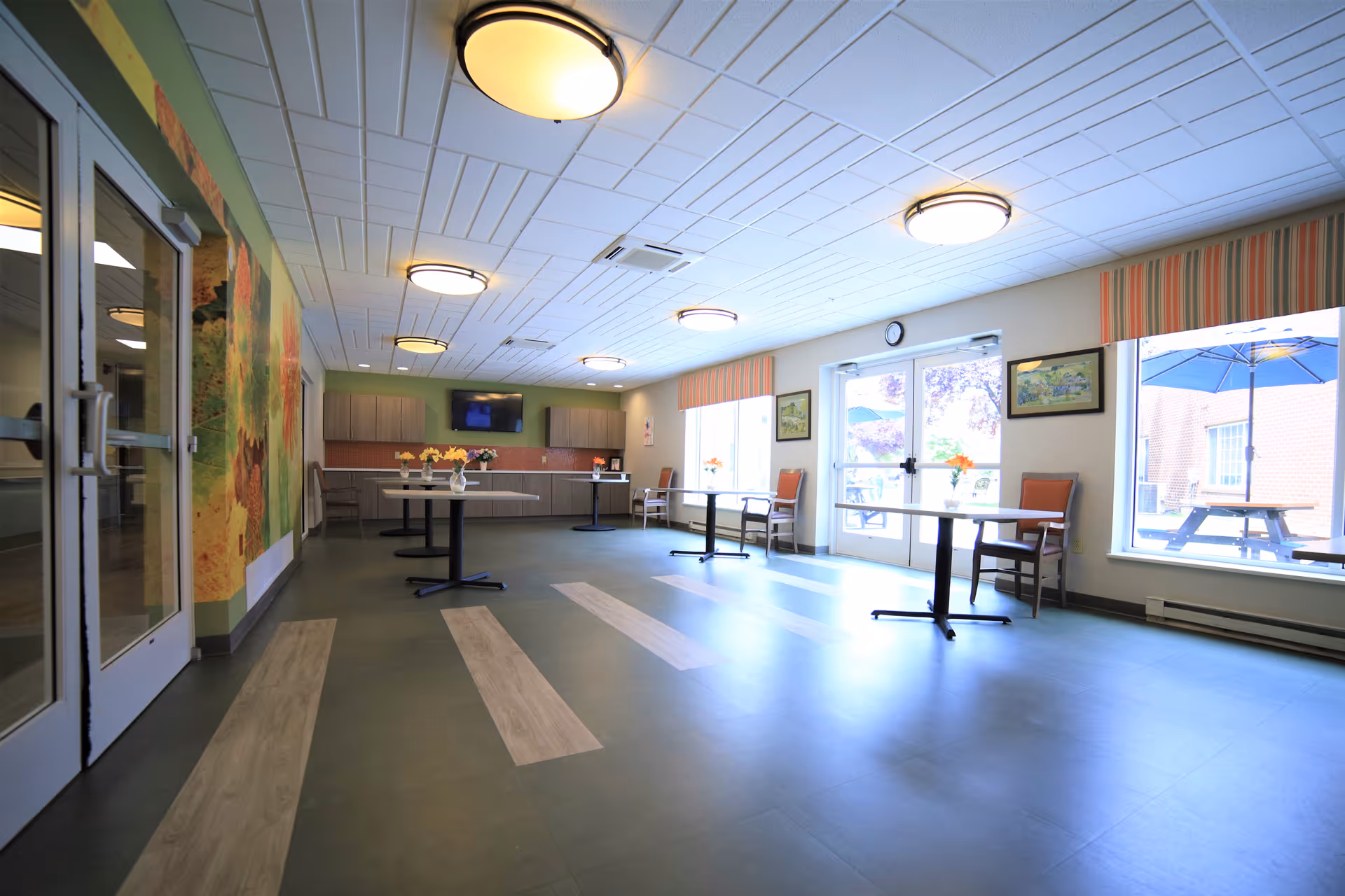 Bright common dining/activity room with tables, chairs, a kitchenette and large windows and doors to an outdoor patio.