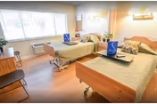 A bright patient bedroom with two hospital-style beds, bedside tables, chairs and a large window with blinds.