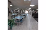 Interior view of a senior living facility dining area with multiple tables and chairs arranged neatly on a tiled floor. The ceiling has recessed lighting, and there are plants and framed pictures on the walls. The space appears clean and well-lit.