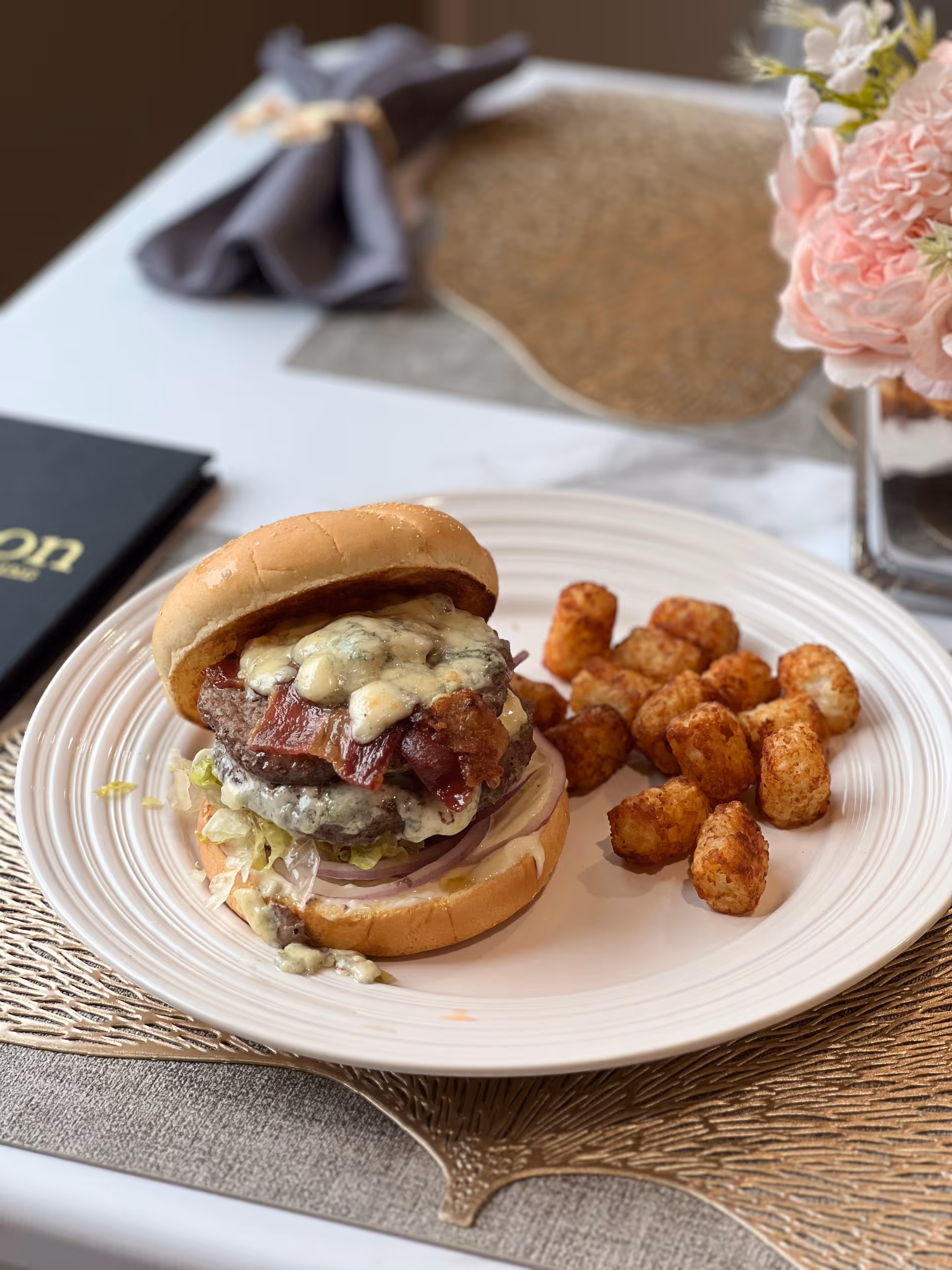 A plate with a cheeseburger topped with bacon, lettuce, and onions, served with a side of tater tots on a white plate. The plate is on a table with a decorative placemat, a folded napkin, a menu, and a vase with pink flowers in the background.