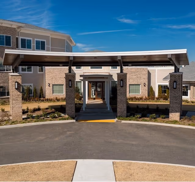 Front entrance of a senior living facility with a covered drop-off area supported by brick pillars, surrounded by landscaped bushes and a clear blue sky.