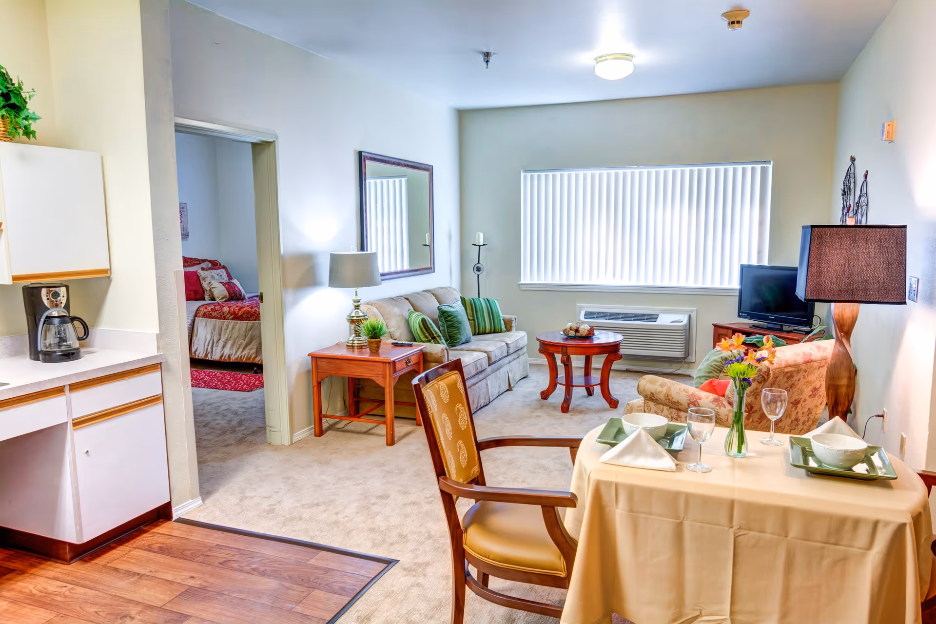 A bright and cozy living area in a senior living facility with a small dining table set for two, a beige sofa with green cushions, a floral armchair, a wooden coffee table, and a TV on a stand. The room has a large window with vertical blinds and a lamp on a side table. To the left, there is a kitchenette with a coffee maker and a doorway leading to a bedroom with a bed covered in red and beige bedding.