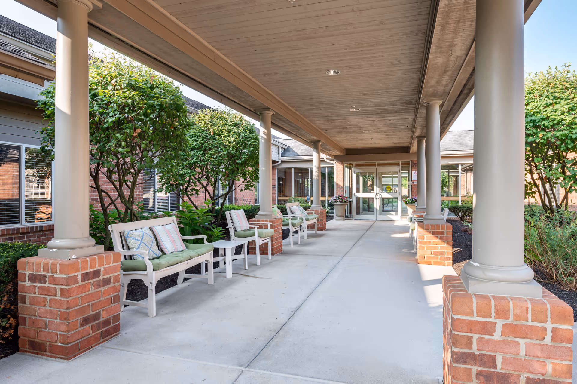 Covered outdoor walkway at Brookdale Pinnacle with white benches and chairs featuring green cushions and decorative pillows, brick columns supporting the roof, and neatly trimmed bushes and trees along the sides.