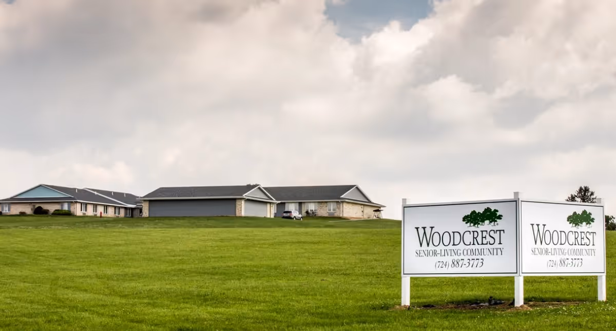 Wide view of a senior living community building with a large green lawn in the foreground under a cloudy sky. Two white signs with green tree logos display the name 'Woodcrest Senior Living Community' and a phone number.