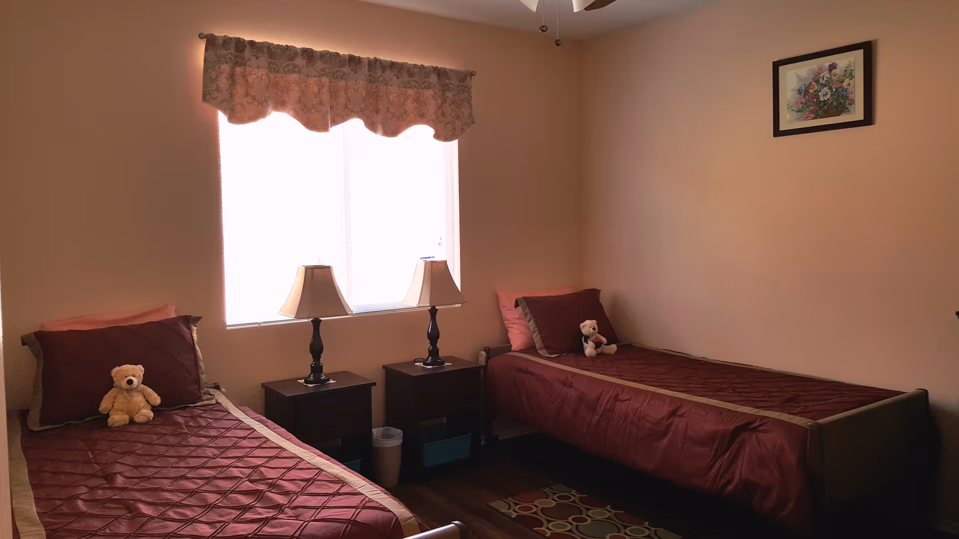 A small bedroom with two single beds placed parallel to each other, each with a maroon bedspread and a teddy bear on top. Between the beds are two dark wooden nightstands, each with a lamp. A window with a floral valance is centered on the wall behind the nightstands. A framed floral picture hangs on the adjacent wall. The room has beige walls and a ceiling fan with lights.