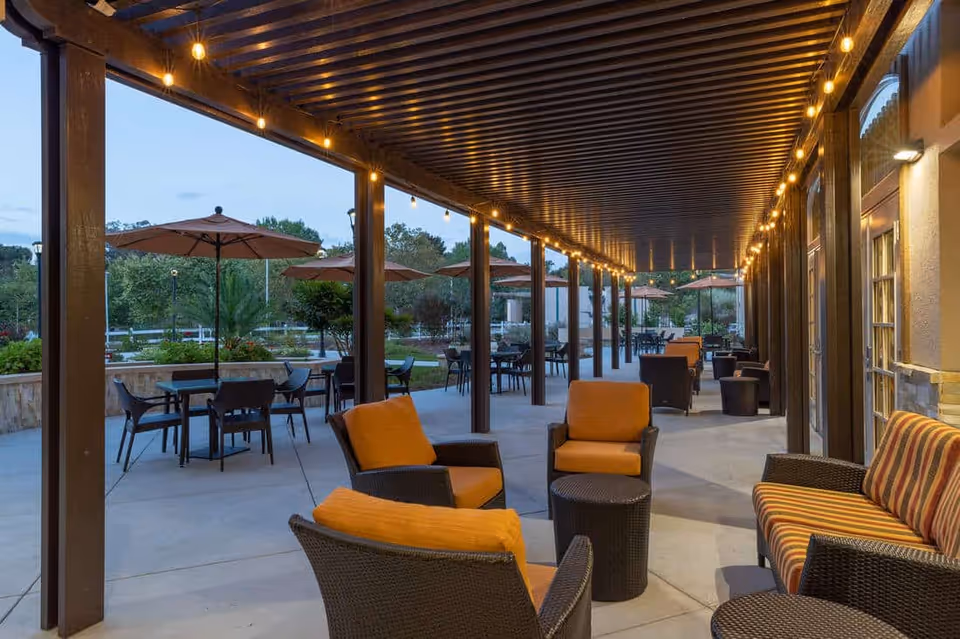 Covered outdoor patio with wicker seating and orange cushions, dining tables with umbrellas, and string lights.