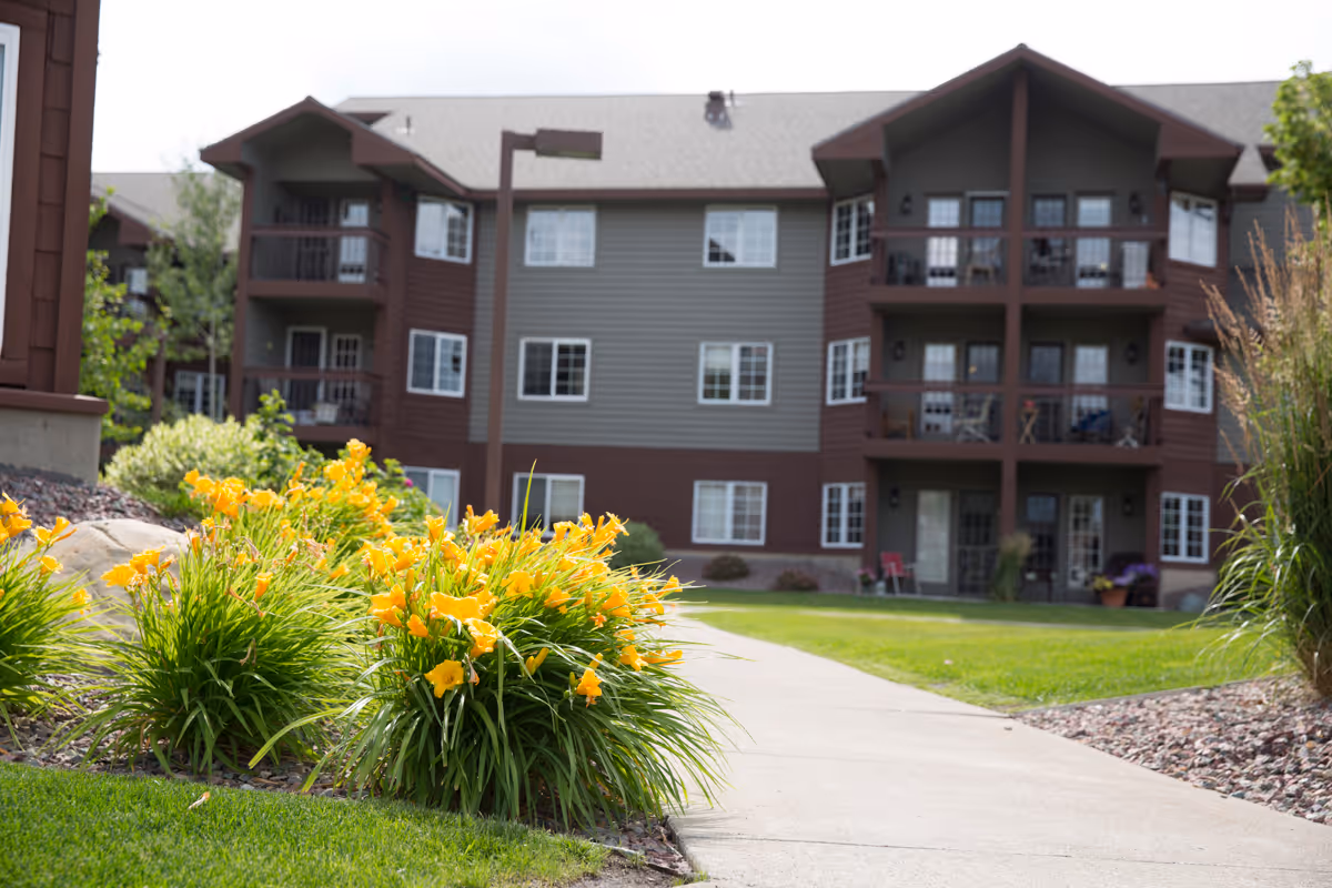Outdoor view of a senior living facility building with balconies, surrounded by green grass, yellow flowers, and a concrete walkway leading towards the entrance.