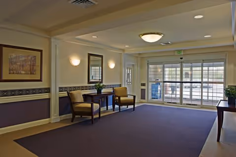 A spacious lobby area with a large dark carpet, two wooden chairs with cushions, a wooden table with a plant, a wall mirror, framed artwork, and glass double doors leading outside.