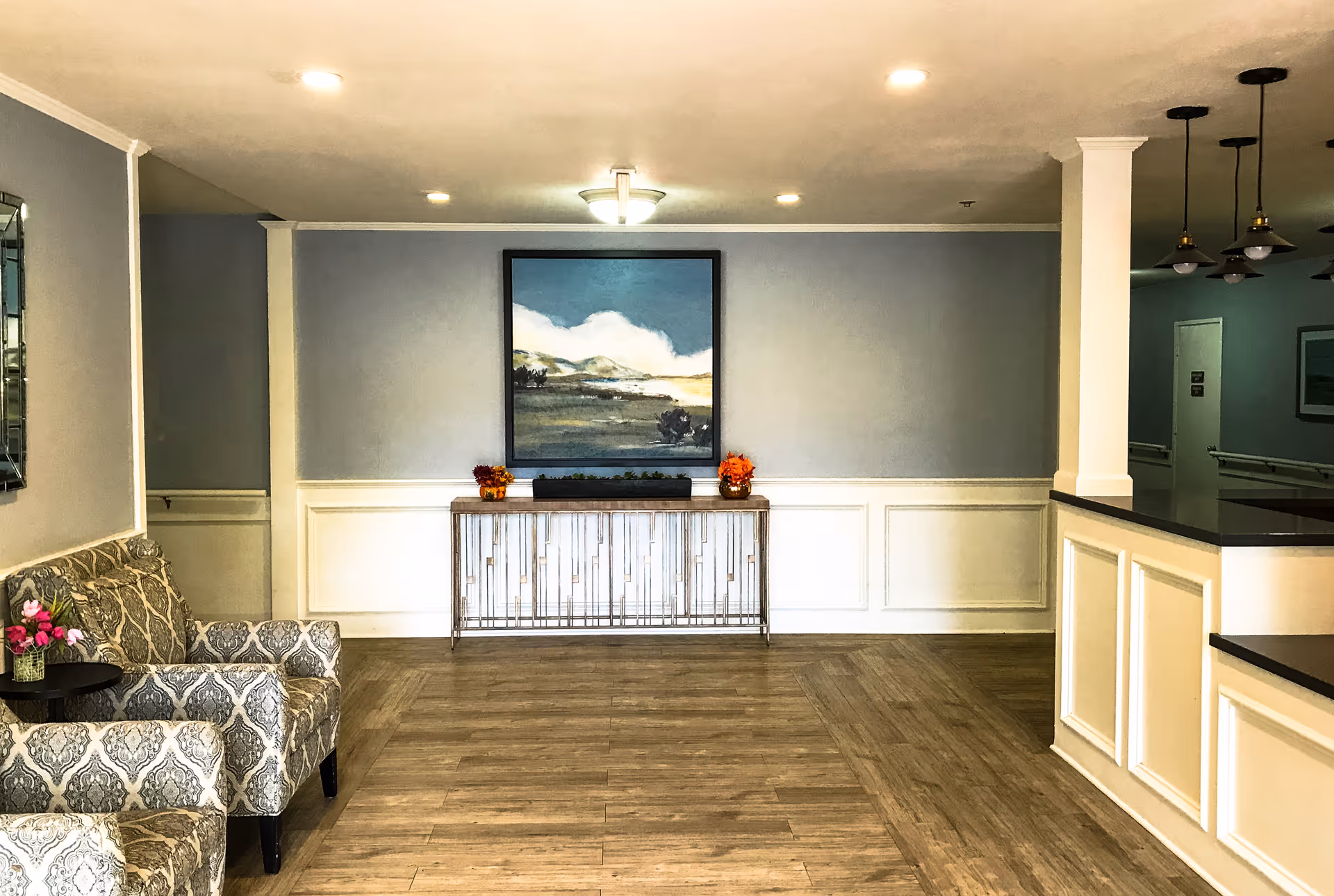 A well-lit interior common area with patterned armchairs on the left, a decorative console table against a light blue wall with white wainscoting, and a large framed landscape painting above the table. The floor is wood, and there is a counter with pendant lights hanging above it on the right side.