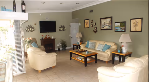 A bright living room with cream leather sofas and chairs around a wooden coffee table, a wall-mounted TV above a mantel, framed artwork, lamps, and tiled floors.