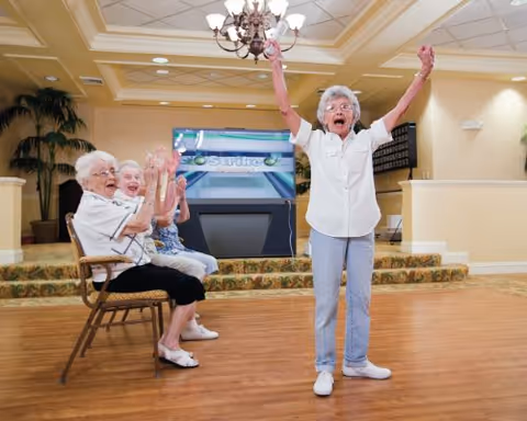 An elderly woman stands with her arms raised in excitement in a spacious room with wooden floors and a chandelier. Three other elderly women sit on chairs nearby, clapping and smiling. The room has warm lighting, plants, and a large screen in the background.
