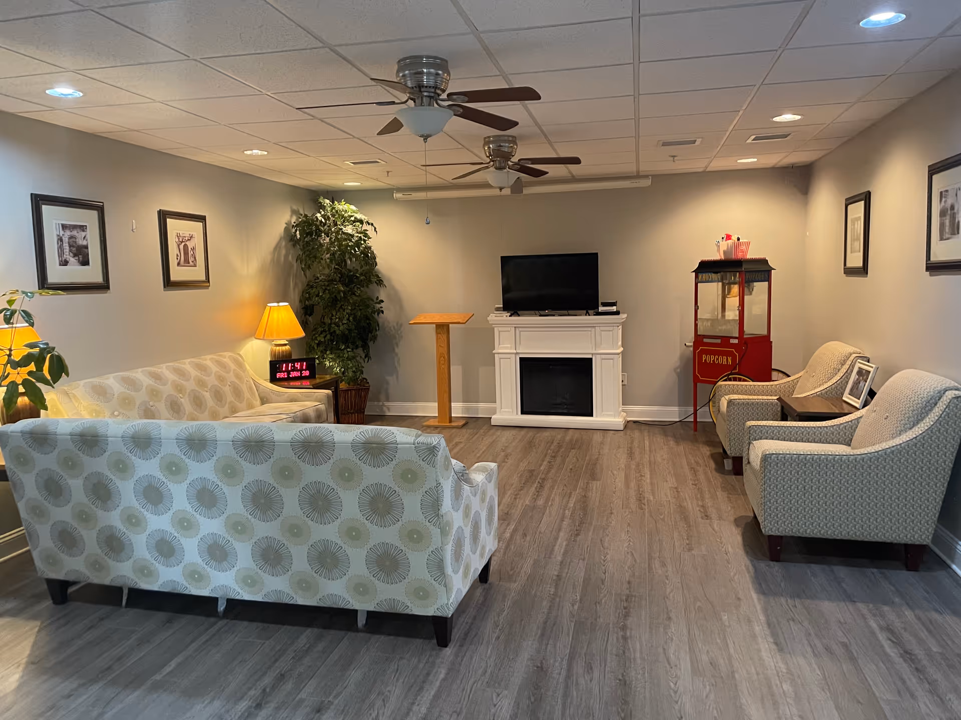 A cozy living room area with two patterned sofas and two armchairs arranged around a TV on a white fireplace mantel. There is a red popcorn machine in the corner, a wooden podium, a potted plant, framed pictures on the walls, and ceiling fans overhead. The room has wood flooring and soft lighting from lamps and recessed ceiling lights.
