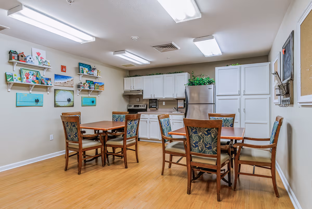 Communal dining room with two wooden tables and chairs in front of a kitchenette with white cabinets and a stainless refrigerator.