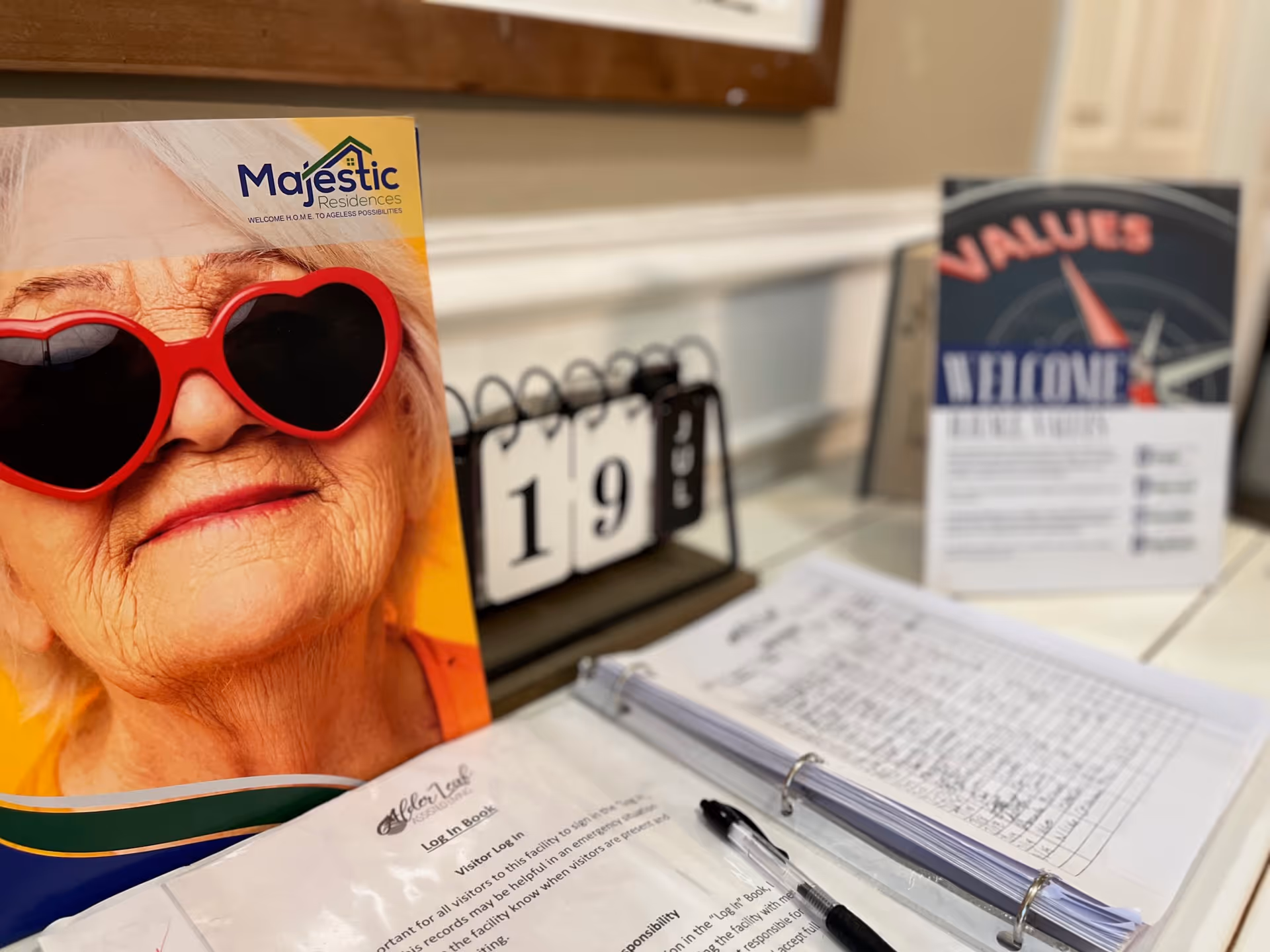 A reception table with a brochure featuring an elderly woman in heart-shaped sunglasses, a flip calendar showing '19', and an open sign-in binder with a pen.