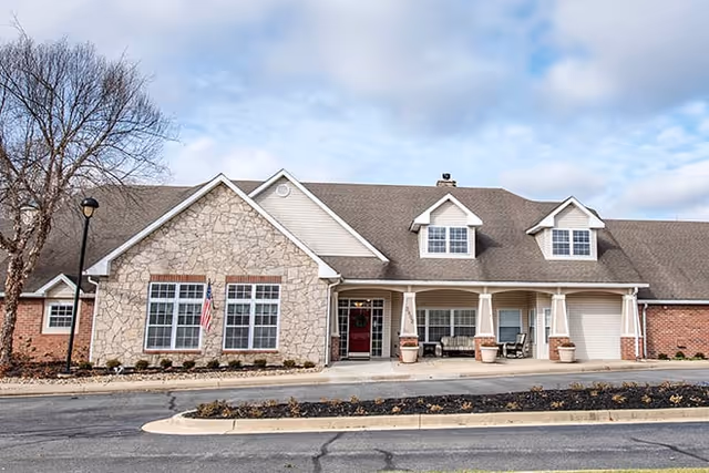 Front exterior of a single-story senior living building with a stone facade, covered porch with columns, and dormer windows under a partly cloudy sky.
