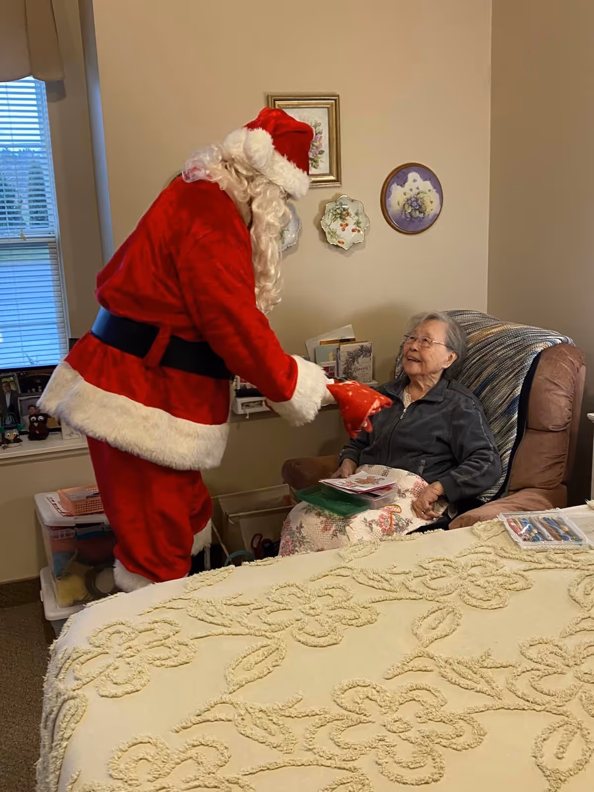 An elderly woman sitting in a recliner chair covered with a floral blanket, smiling and looking at a person dressed as Santa Claus who is handing her a red gift bag in a cozy room with framed wall decorations and a window with blinds.