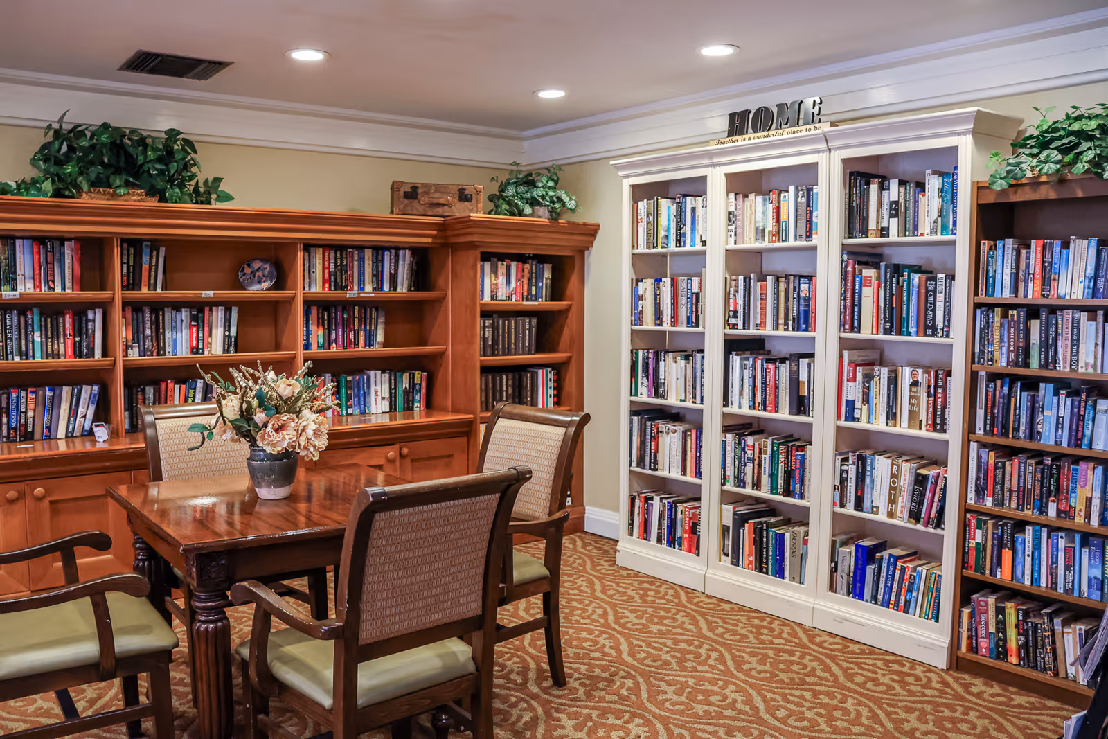 A cozy library room with wooden bookshelves filled with books, a wooden table with a flower vase in the center, and four cushioned chairs around the table. The room has carpeted flooring with a patterned design and decorative plants on top of the bookshelves. A decorative sign on top of a white bookshelf reads 'HOME'.
