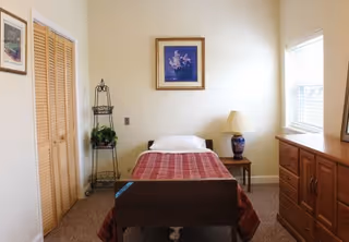 A small, simple bedroom with a single bed covered with a red patterned blanket. There is a wooden dresser with drawers on the right side under a window with blinds. A nightstand with a decorative lamp is next to the bed. On the left side, there is a closet with wooden louvered doors and a metal plant stand with a potted plant. A framed picture hangs on the wall above the bed.