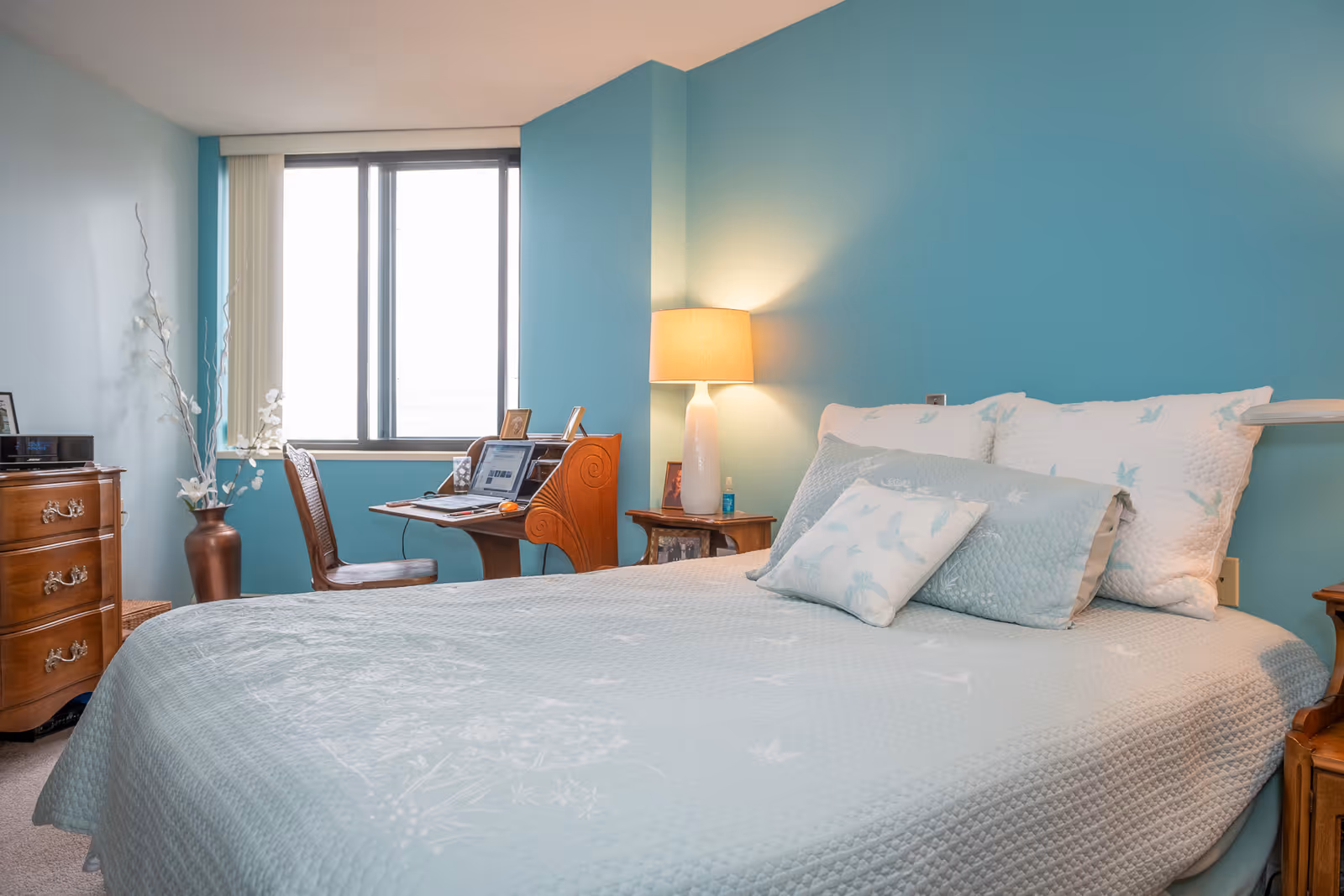 Light-blue bed with coordinating pillows in a teal-painted bedroom with a bedside lamp and a desk by the window.