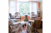 A cozy living room with floral upholstered armchairs, a small round table with picture frames in front of a large window with sheer white curtains, and additional wooden chairs arranged around the space. The room has soft peach-colored walls and a patterned rug on the floor.