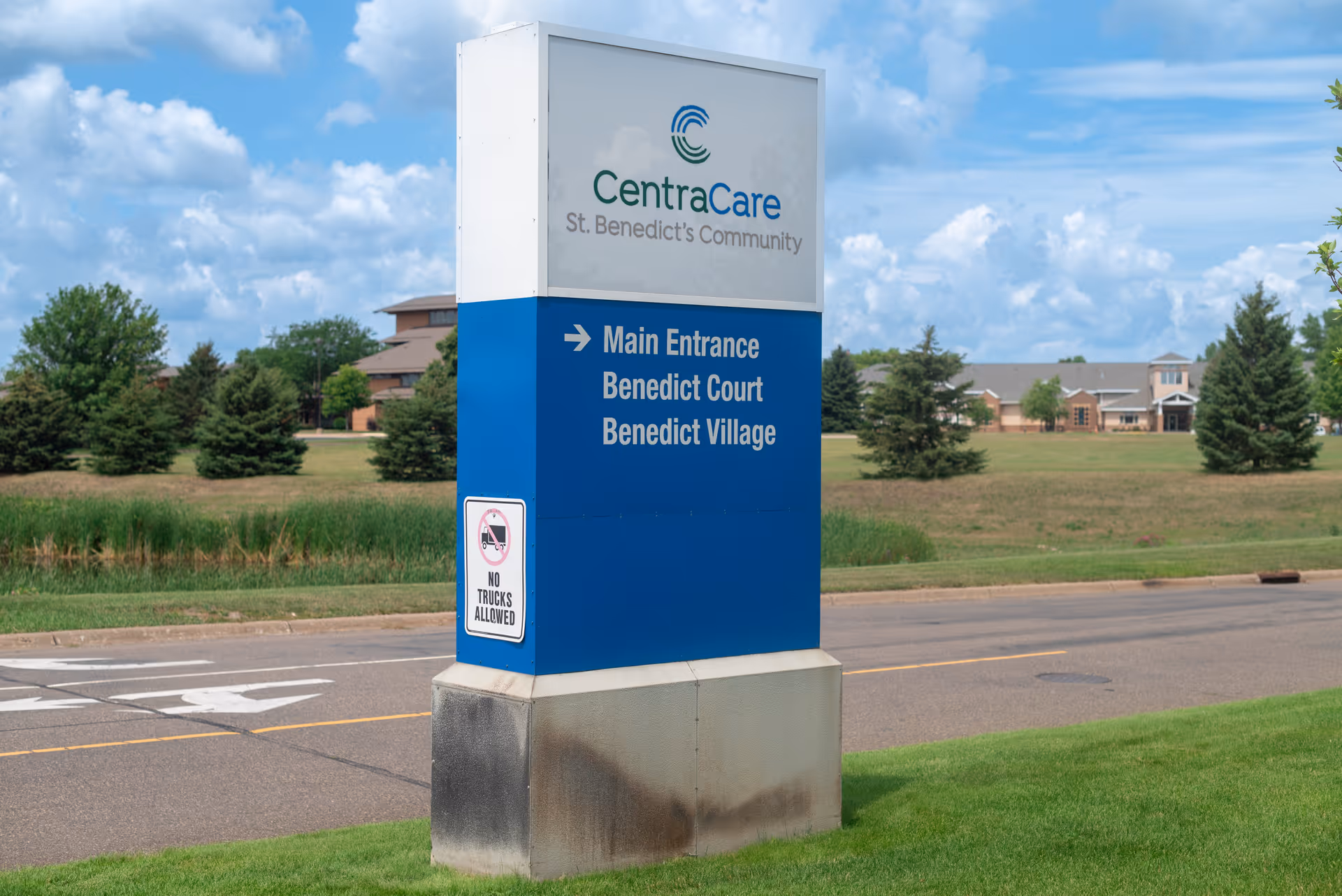 Outdoor image showing a large blue and white sign for CentraCare St. Benedict's Community with directions to the Main Entrance, Benedict Court, and Benedict Village. The sign is positioned on a grassy area beside a road with trees and buildings in the background under a partly cloudy sky.