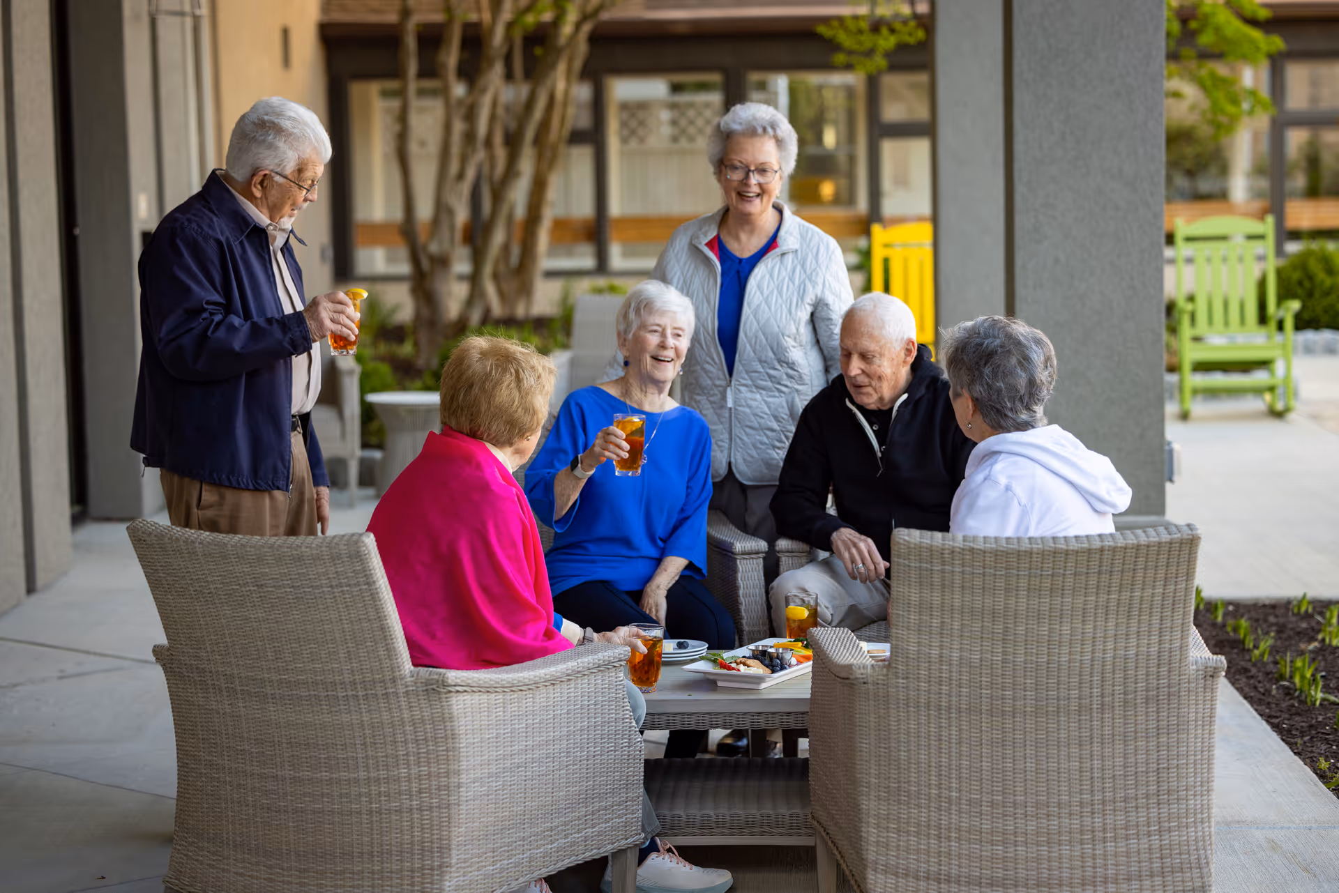 A group of six elderly people socializing outdoors on a patio. Four are seated on wicker chairs around a small table with drinks and snacks, while two stand nearby. They appear to be enjoying a pleasant conversation in a garden-like setting with greenery and colorful chairs in the background.