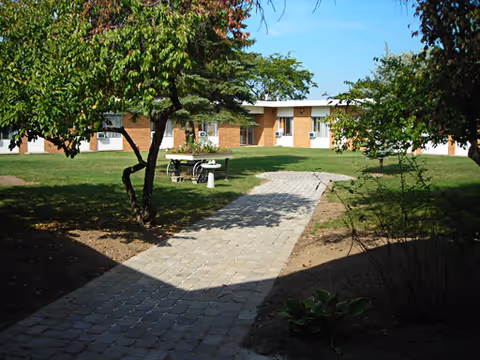 A paved walkway leading through a grassy courtyard with trees and shrubs, surrounded by a single-story brick building under a clear blue sky.