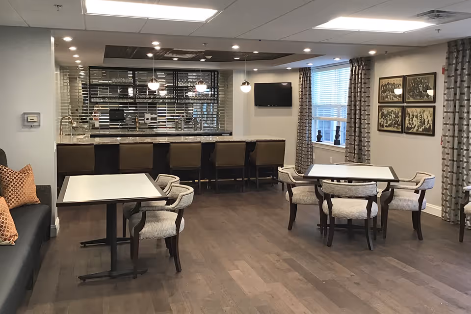 A modern dining area with several tables and cushioned chairs, a long counter with bar stools, pendant lights hanging from the ceiling, a wall-mounted TV, and windows with patterned curtains. The room has wooden flooring and framed pictures on the wall.