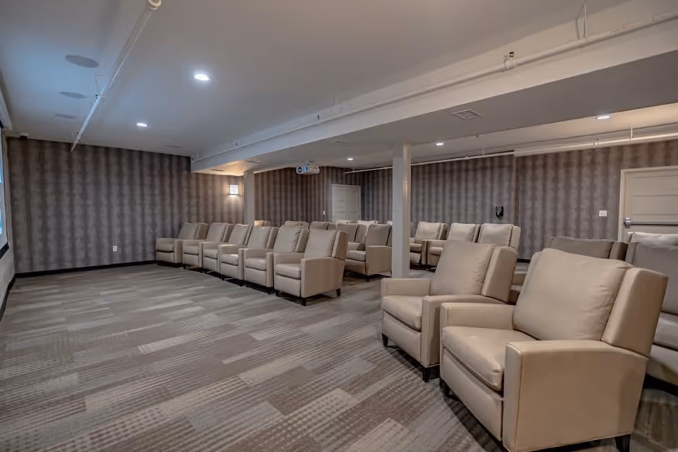 Carpeted screening room with rows of beige reclining chairs facing a projector screen in a senior living facility.