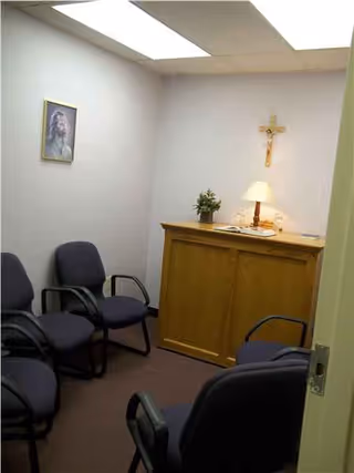 A small chapel-like room with a wooden cabinet, crucifix, lamp, plant, and several blue chairs.