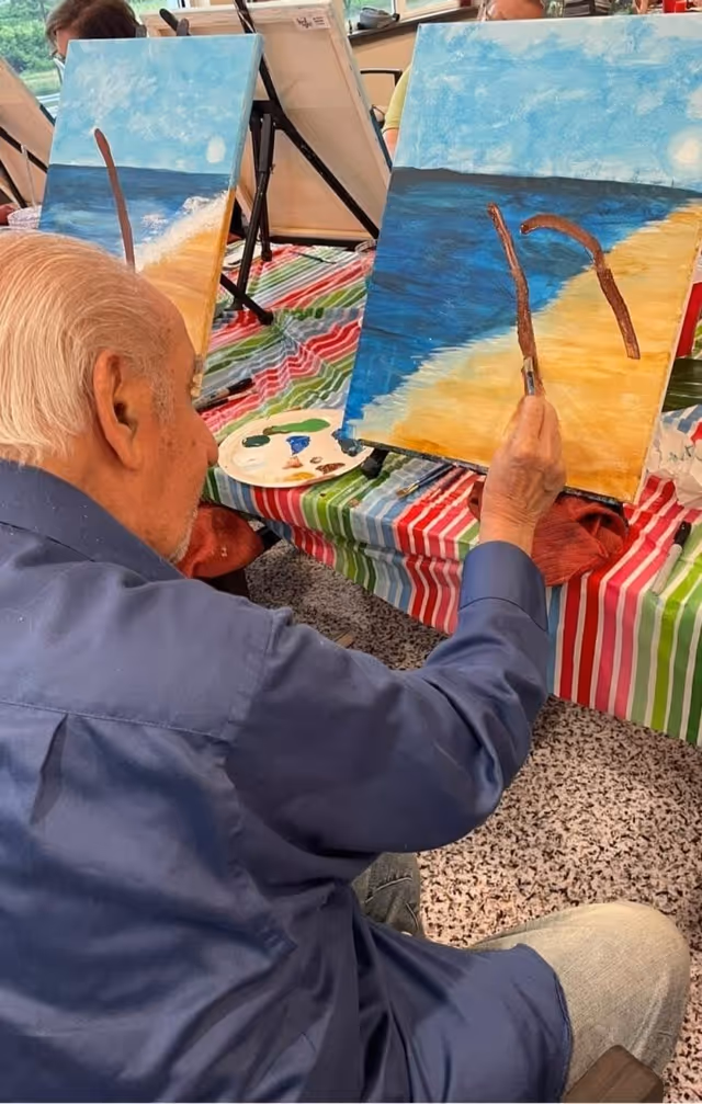 An elderly man in a blue shirt is painting a beach scene on a canvas set on an easel. The painting depicts a sandy shore with blue ocean water and a sky with clouds. The man is holding a paintbrush and adding brown details to the painting. The table is covered with a colorful striped tablecloth, and there are other canvases and painting supplies visible in the background.