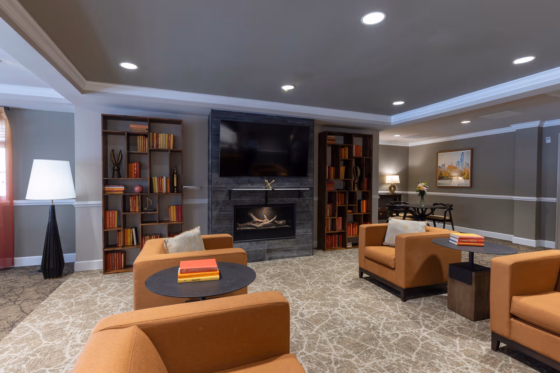 A cozy living room area with four orange armchairs arranged around two small round tables, each holding a few books. The room features a modern fireplace with a large flat-screen TV mounted above it, flanked by two tall bookshelves filled with books and decorative items. The walls are painted gray with white trim, and the carpet has a light, abstract pattern. A tall floor lamp stands near a window with sheer curtains, and a small table with a lamp and flowers is visible in the background.