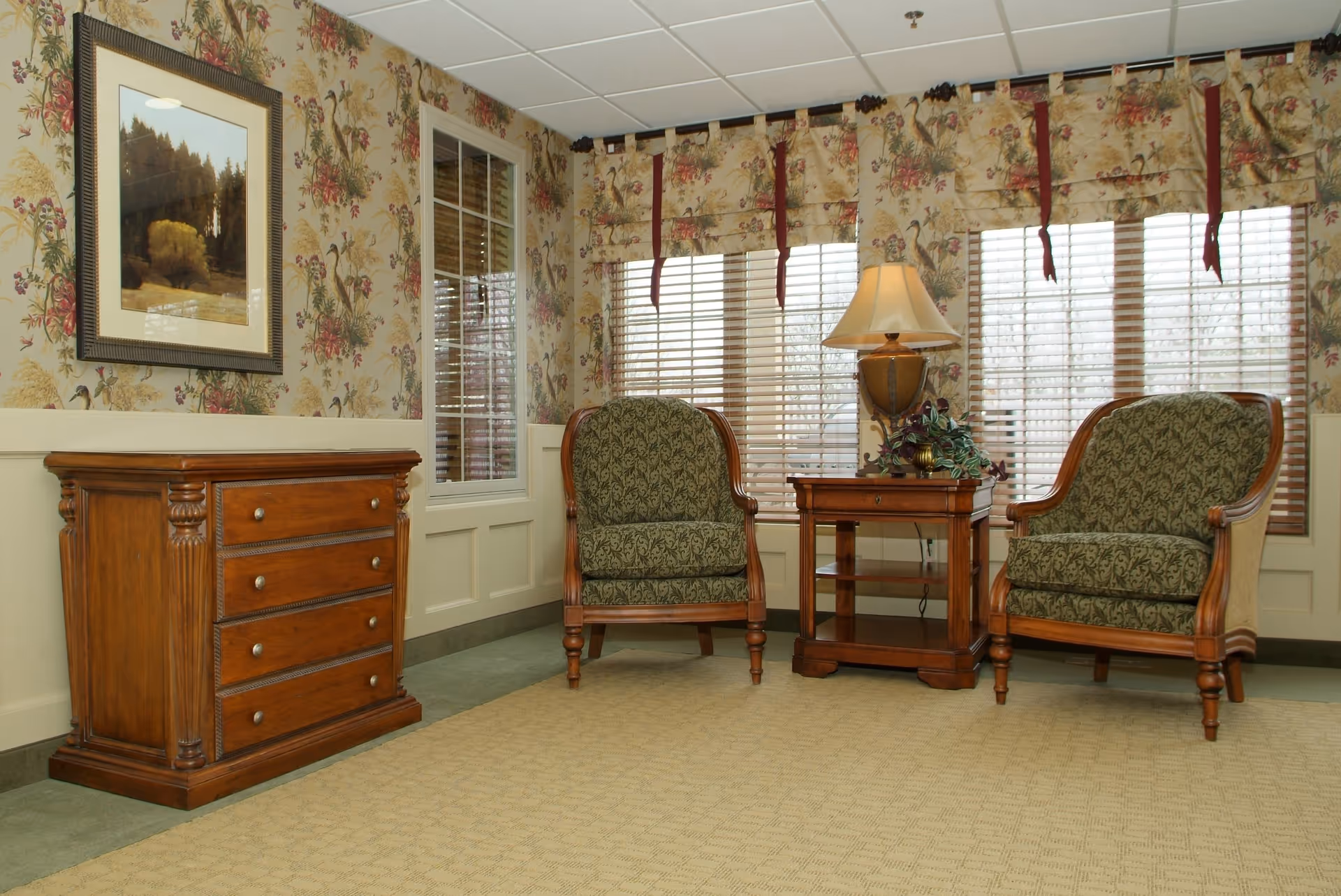 A cozy sitting area in a senior living community with two green patterned armchairs, a wooden side table with a lamp and a plant, floral wallpaper, a framed landscape painting, and windows with wooden blinds and floral valances.