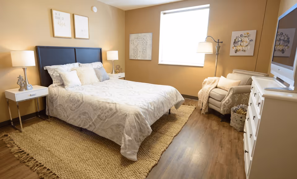 A cozy bedroom with a large bed covered in a light patterned comforter, two white nightstands with lamps on either side, a beige armchair with a throw blanket, a white dresser with a TV on top, and a window letting in natural light. The walls are painted a warm beige color, and there are framed artworks hanging on the walls.