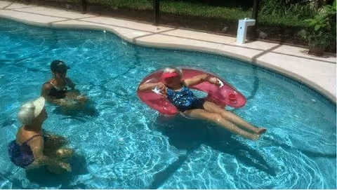 Three elderly women enjoying a sunny day in a swimming pool. One woman is floating on a pink inflatable ring, while the other two are standing in the water nearby. The pool is surrounded by a concrete deck and some greenery.