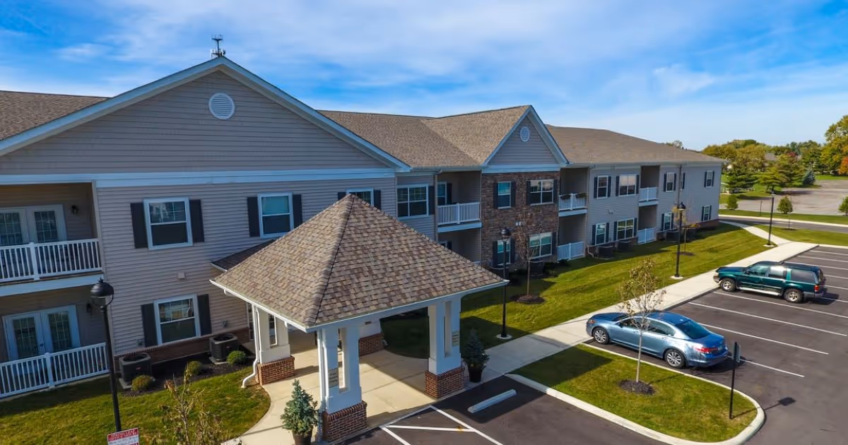 Two-story senior living building with a covered entrance portico, balconies, landscaped lawn, and a parking lot with parked cars.
