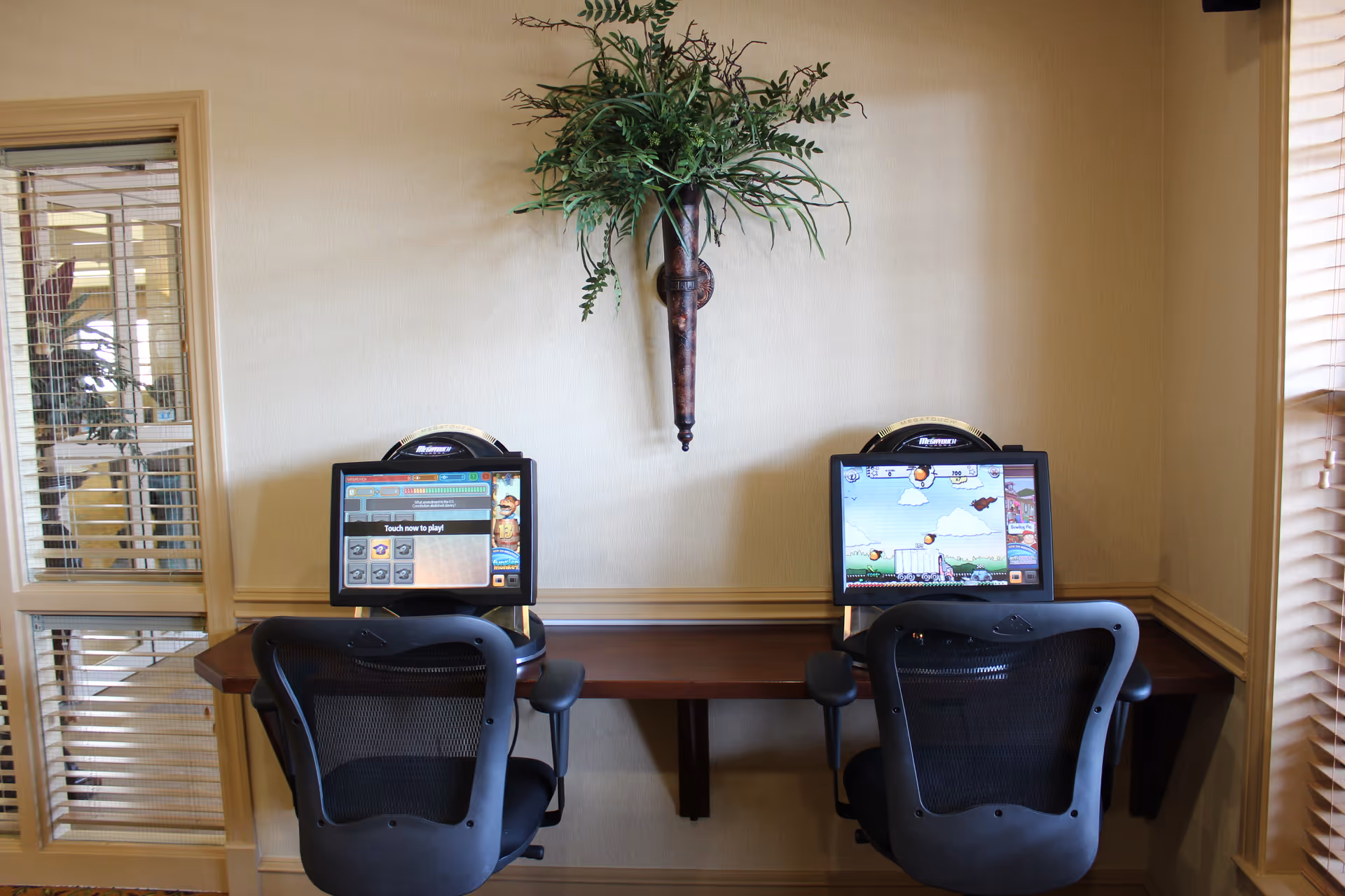 Two computer stations with black mesh office chairs in front of a wooden desk against a beige wall. Each computer screen displays a game interface. Above the desk is a decorative wall-mounted plant. To the left is a window with blinds and a view into another room.