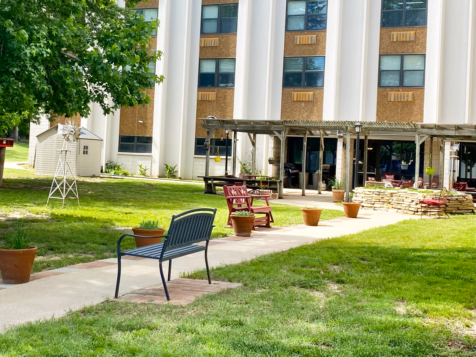 Outdoor seating area at Arkansas City Presbyterian Manor with benches, chairs, potted plants, a small windmill decoration, and a pergola attached to the building. The building has multiple windows and a grassy lawn surrounds the paved walkway.