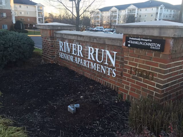 Brick sign at the entrance of River Run Senior Apartments with the address number 13900 and a plaque indicating professional management by The Franklin Johnston Group. Apartment buildings and parked cars are visible in the background.