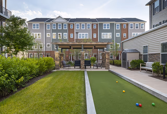Outdoor courtyard area of a senior living facility featuring a bocce ball court with balls on the green surface, a grassy lawn, a shaded seating area with chairs under a pergola supported by stone pillars, surrounded by multi-story residential buildings and landscaping.