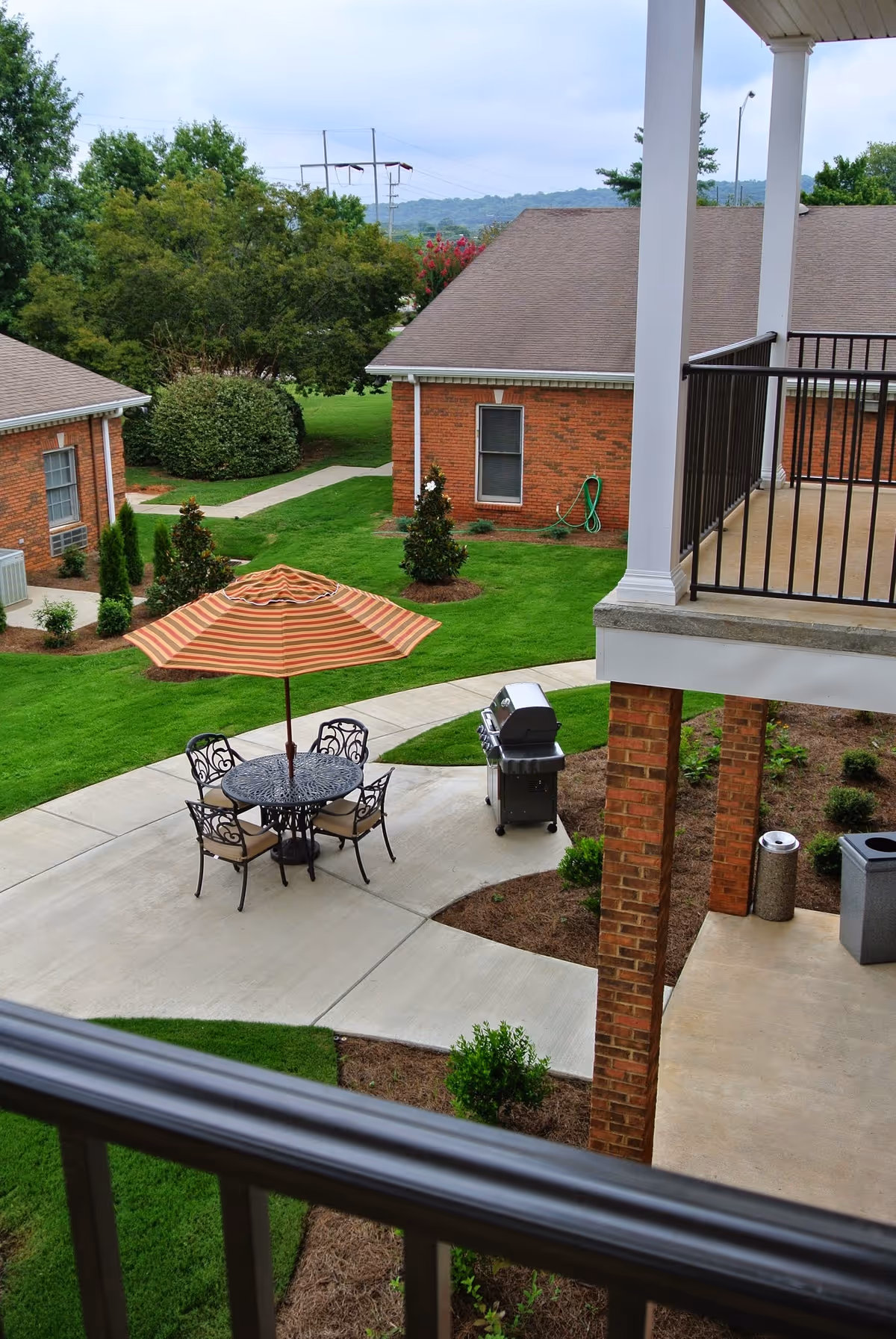 Outdoor patio area in a senior living community with a round metal table, four chairs, and a striped umbrella. There is a barbecue grill nearby on a concrete pathway surrounded by well-maintained green grass and small trees. Brick buildings and a balcony with white columns and black railings are visible.