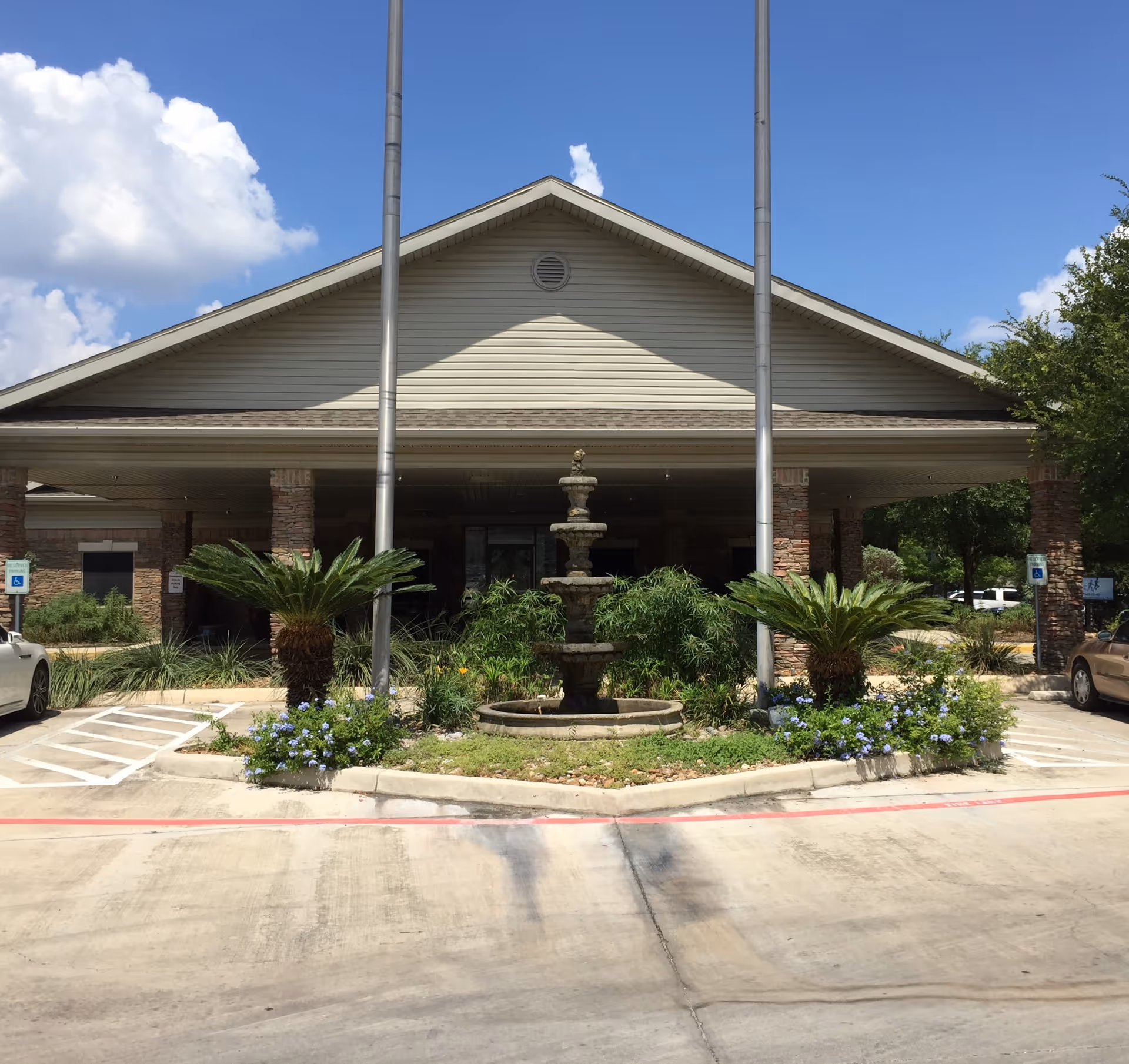 Front exterior view of Silver Tree Nursing and Rehabilitation facility with a covered entrance, two flagpoles, a three-tiered stone fountain, palm plants, and a clear blue sky with some clouds.