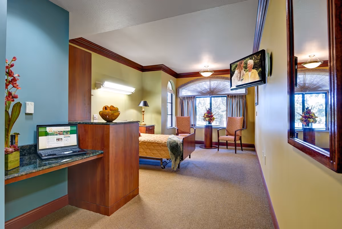 A well-lit senior living bedroom with a single bed covered in a yellow quilt, two upholstered chairs around a small round table with a flower arrangement, a wall-mounted TV showing two elderly people, a large mirror on the wall, and a desk with a laptop and a decorative plant.
