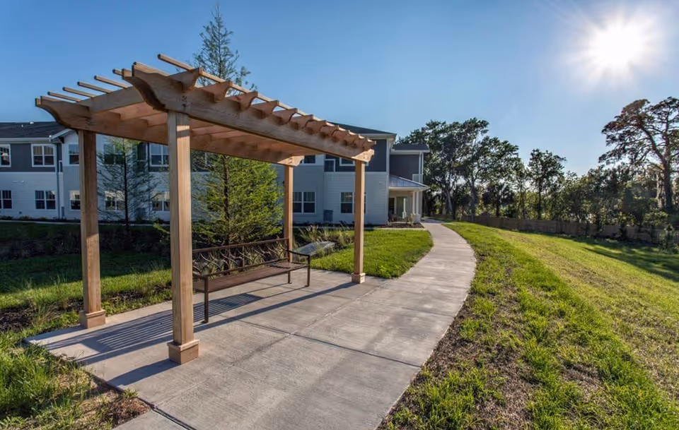 Outdoor area at The Crossings at Riverview featuring a wooden pergola with a bench underneath, a concrete pathway curving alongside a grassy lawn, and a multi-story building in the background under a clear blue sky with the sun shining brightly.