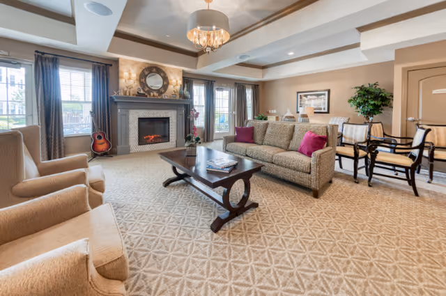 A spacious and well-lit living room with beige patterned carpet, a gray fireplace with a round mirror above it, two large windows with dark curtains, a beige sofa with purple cushions, two beige armchairs, a dark wooden coffee table with magazines, a guitar leaning against the wall, and a dining area with chairs and a large plant in the background.