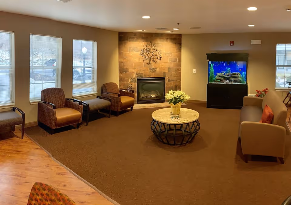 A cozy living room area with brown carpet and beige walls featuring a stone fireplace with a metal tree decoration above it. There are multiple chairs and a sofa arranged around a round coffee table with a potted plant on it. A large aquarium is visible against the far wall, and several windows let in natural light.