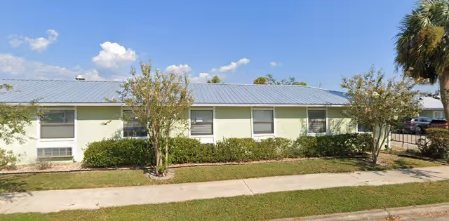 Single-story light-green building with a metal roof, row of windows, shrubs, a sidewalk and palm trees under a blue sky.