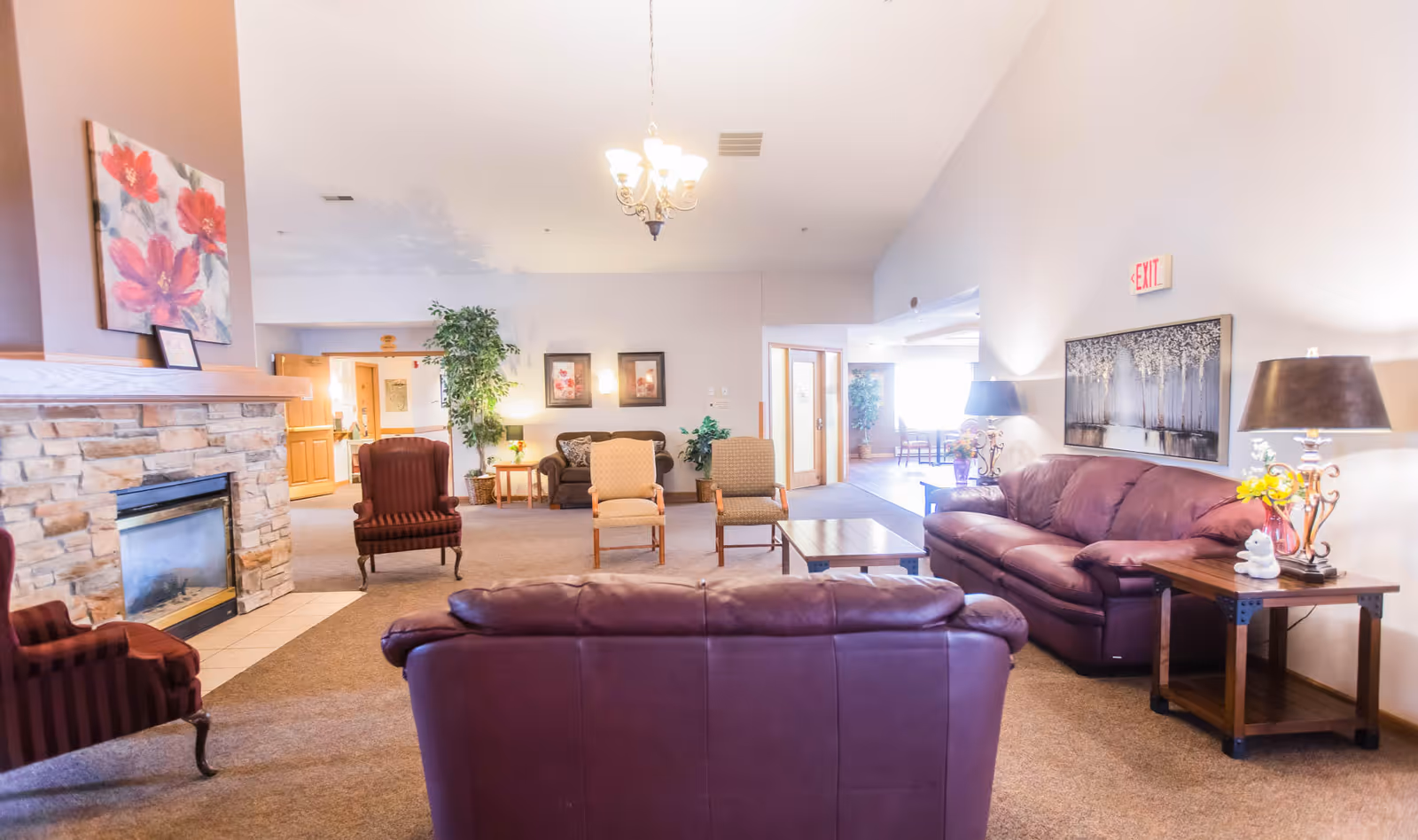 A spacious and well-lit living room area in an assisted living facility featuring multiple seating options including two purple leather sofas, two striped armchairs, and two wooden chairs with cushions. The room has a stone fireplace on the left wall with a floral painting above it, a chandelier hanging from the ceiling, and various plants and framed pictures decorating the space. There are side tables with lamps and flowers, and an exit sign is visible on the right wall.